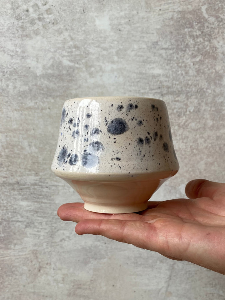 A handmade ceramic cup with a beige base and dark blue-gray speckled glaze, held in a person&