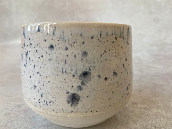 A handmade ceramic bowl with a speckled glaze, featuring a cream-colored base with dark blue-gray and black flecks, photographed on a neutral textured surface.