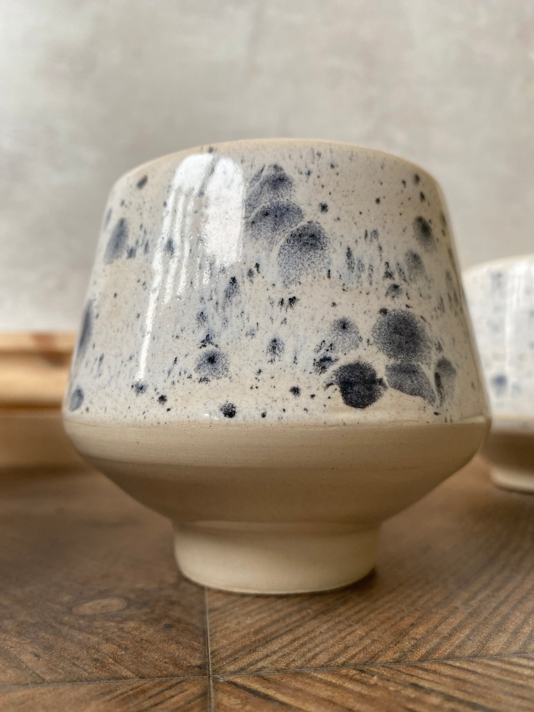 A handmade ceramic cup with a conical shape, featuring a glossy speckled cream glaze on the upper half and a matte beige base, set on a wooden surface.