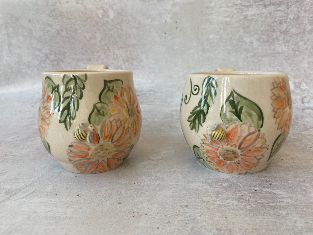 Two small ceramic cups with a cream-colored glaze, decorated with orange flowers, green leaves, and yellow-and-black bees, set against a textured gray background.