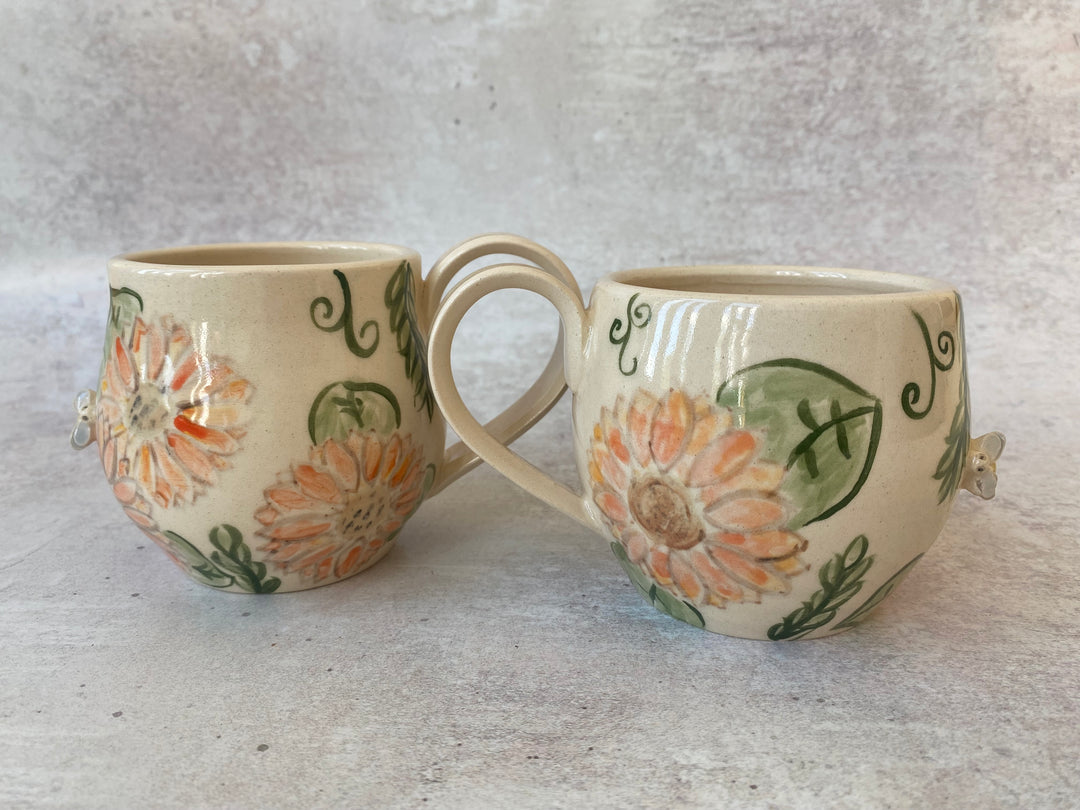 Two small ceramic mugs with floral hand-painted designs in orange, green, and beige, displayed on a textured gray surface.