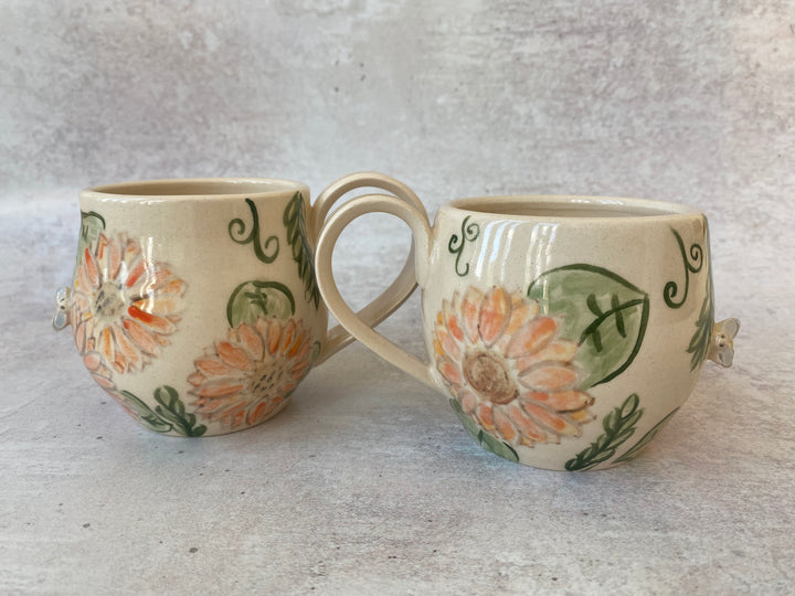 Two small ceramic mugs with floral hand-painted designs in orange, green, and beige, displayed on a textured gray surface.