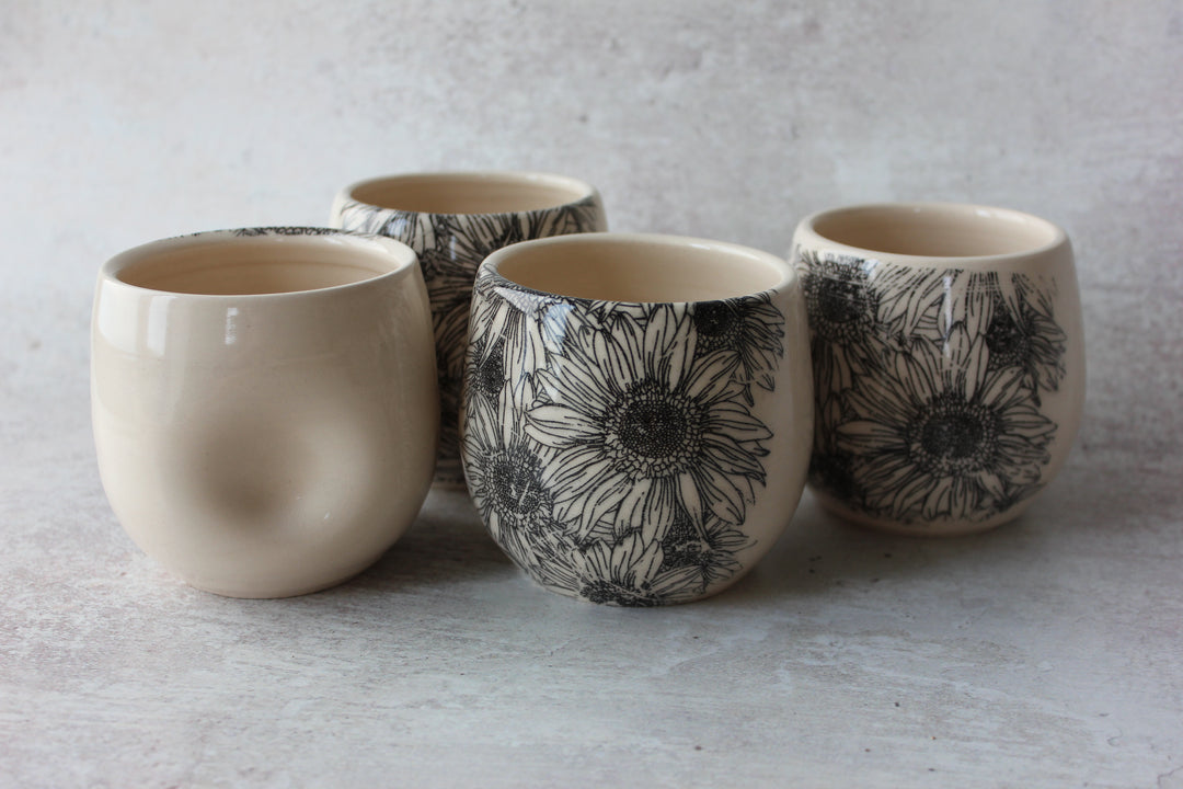 Four small ceramic cups arranged on a light textured surface. Three cups feature a black floral line-art pattern on a warm cream base, and one cup is solid cream with a subtle embossed or sculpted texture.