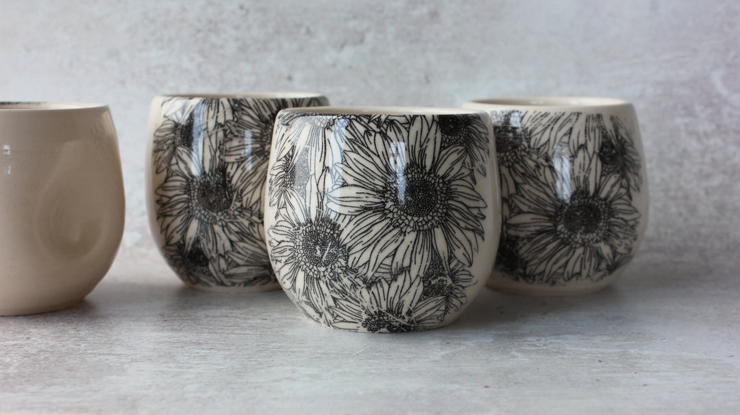 Three small ceramic cups with a black floral sunflower pattern on a cream background, arranged on a light gray surface against a textured gray wall.