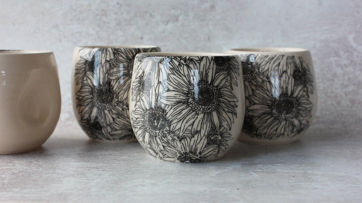 Three small ceramic cups with a black floral sunflower pattern on a cream background, arranged on a light gray surface against a textured gray wall.
