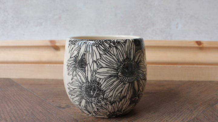 A small ceramic cup with a black floral line-art pattern on a cream background, sitting on a wooden surface against a light wood baseboard and textured wall.
