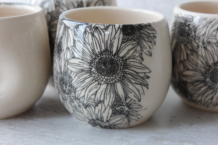 A close-up of a small ceramic cup or tumbler with a black floral line-art print on an off-white background, with other similar cups partially visible in the foreground and background.