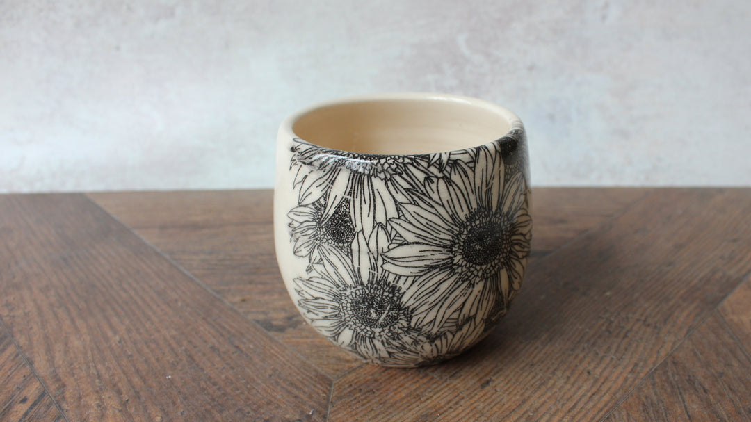 A small ceramic cup with a black floral line-art pattern on a cream-colored exterior, shown on a wooden table against a light gray wall.