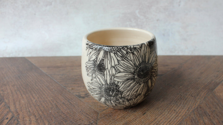 A small ceramic cup with a black floral line-art pattern on a cream-colored exterior, shown on a wooden table against a light gray wall.