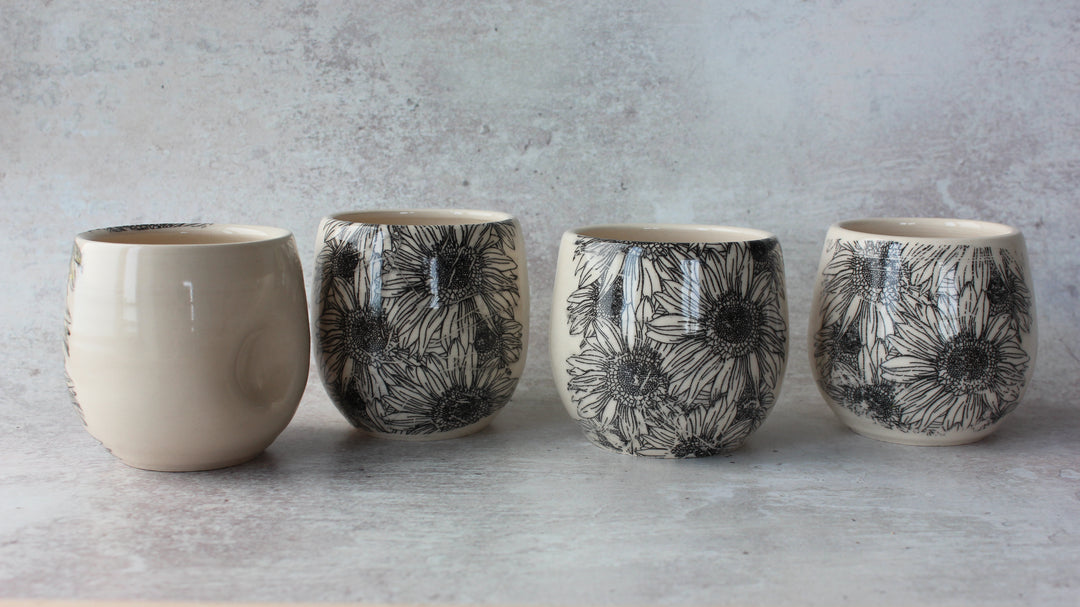 Four ceramic stemless wine glasses arranged in a row against a textured gray background. The first glass is solid cream-colored, while the remaining three feature a black floral line-art pattern on a cream base.