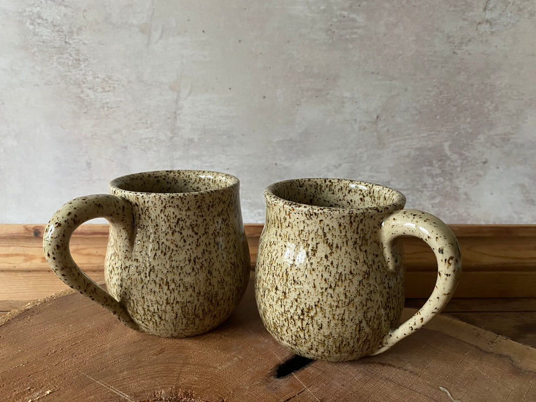 Two speckled ceramic mugs on a wooden surface with a grey concrete wall background.