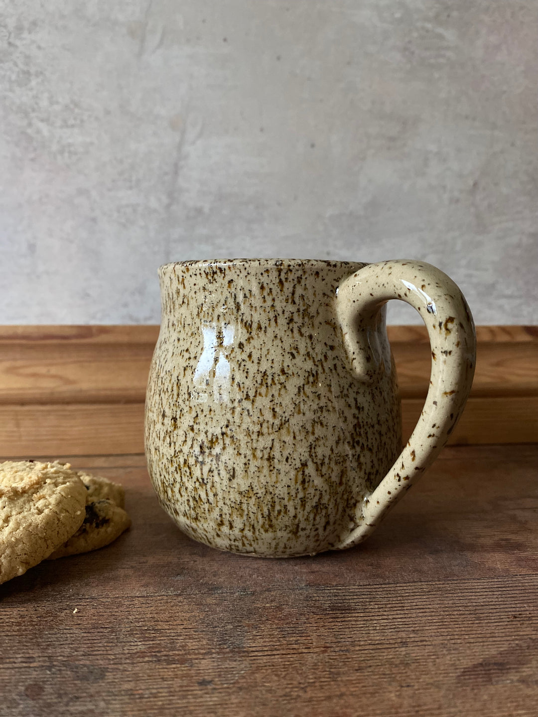 Speckled ceramic mug leaning against a wooden surface with a concrete wall in the background.