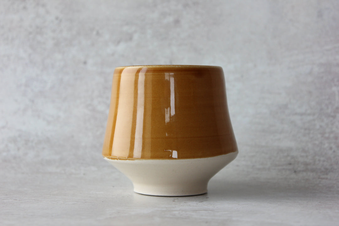 A ceramic cup with a glossy mustard-brown upper body and a matte off-white flared base, set against a textured light gray background.