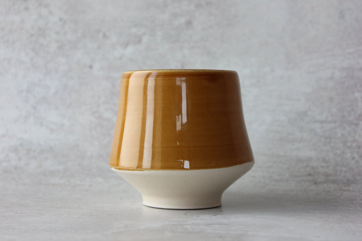 A ceramic cup with a glossy mustard-brown upper body and a matte off-white flared base, set against a textured light gray background.