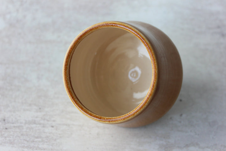 A handmade ceramic cup with a glossy beige interior and a textured brown exterior, photographed from a top-down angle on a light gray stone surface.