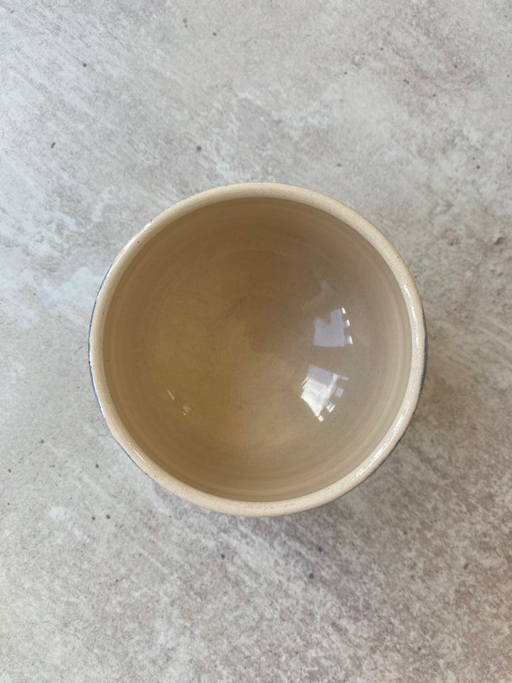 A handmade ceramic bowl with a warm beige interior and a dark blue rim, photographed from a top-down perspective on a textured gray stone surface.