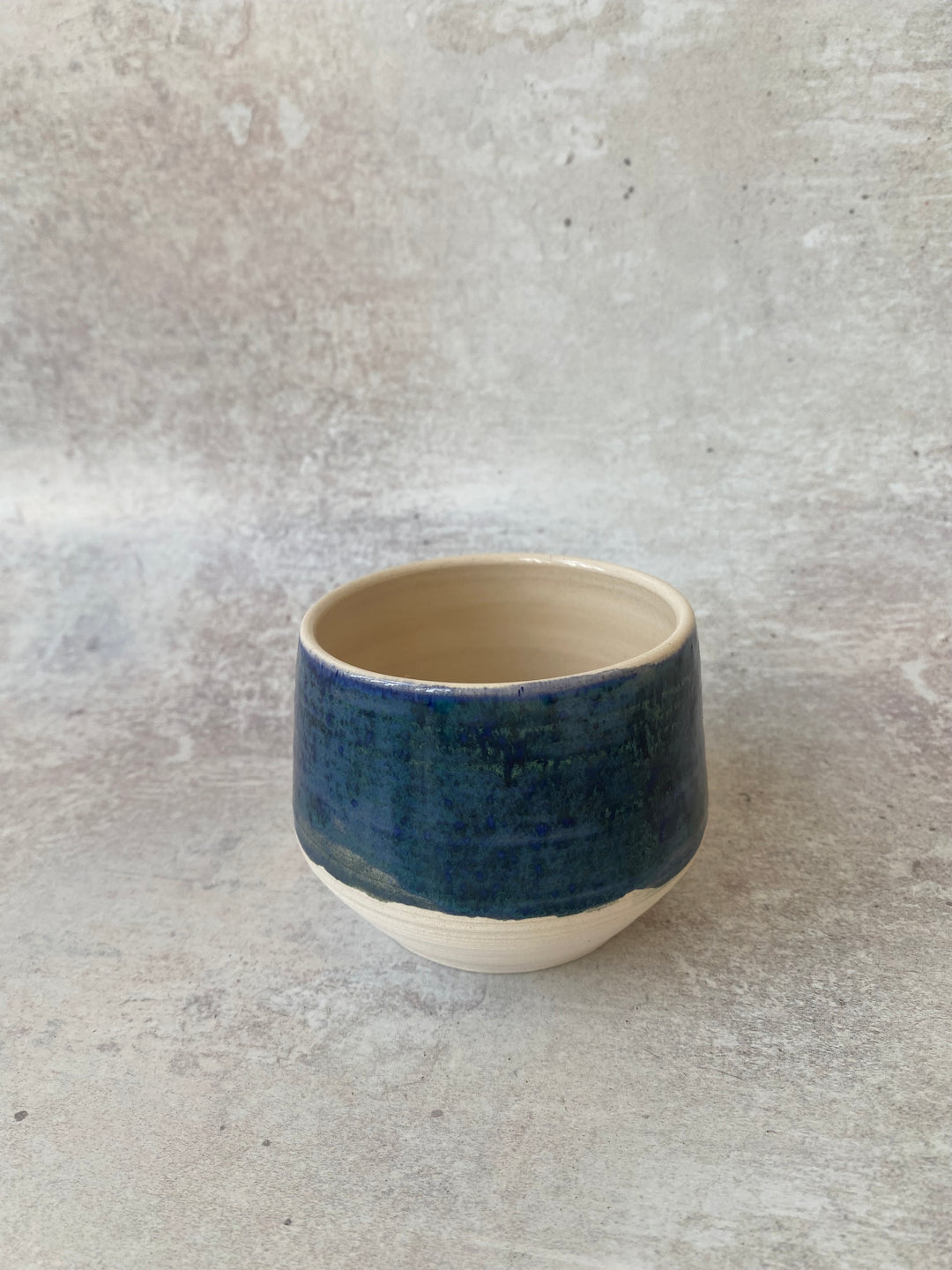 A handmade ceramic cup with a textured dark blue glazed exterior and a natural unglazed beige interior and base, set against a neutral concrete-textured background.