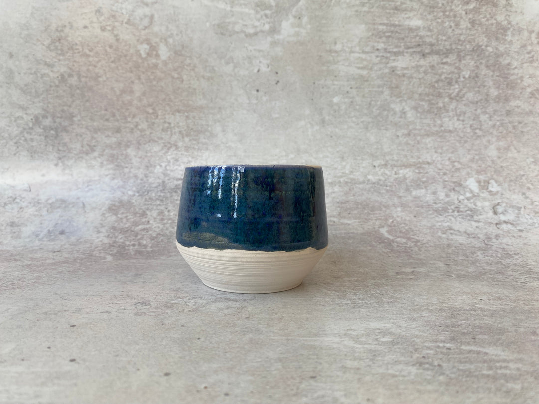 A handmade ceramic cup with a glossy dark blue glazed upper half and a textured, unglazed off-white lower half, set against a neutral gray concrete background.