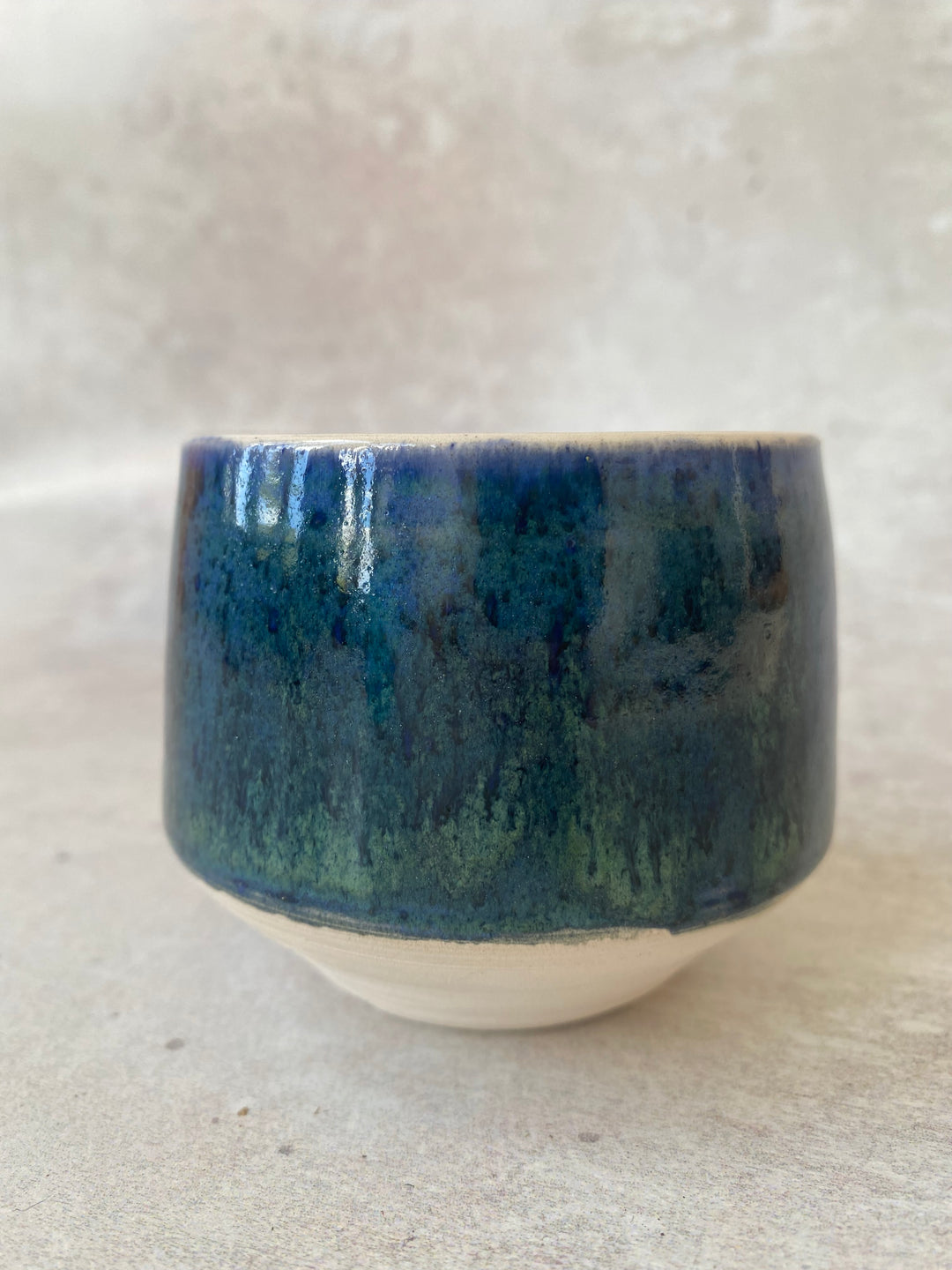 A handmade ceramic cup with a textured, dripping glaze in deep blue and green, featuring a contrasting unglazed, natural beige clay base, set against a neutral, light beige textured background.