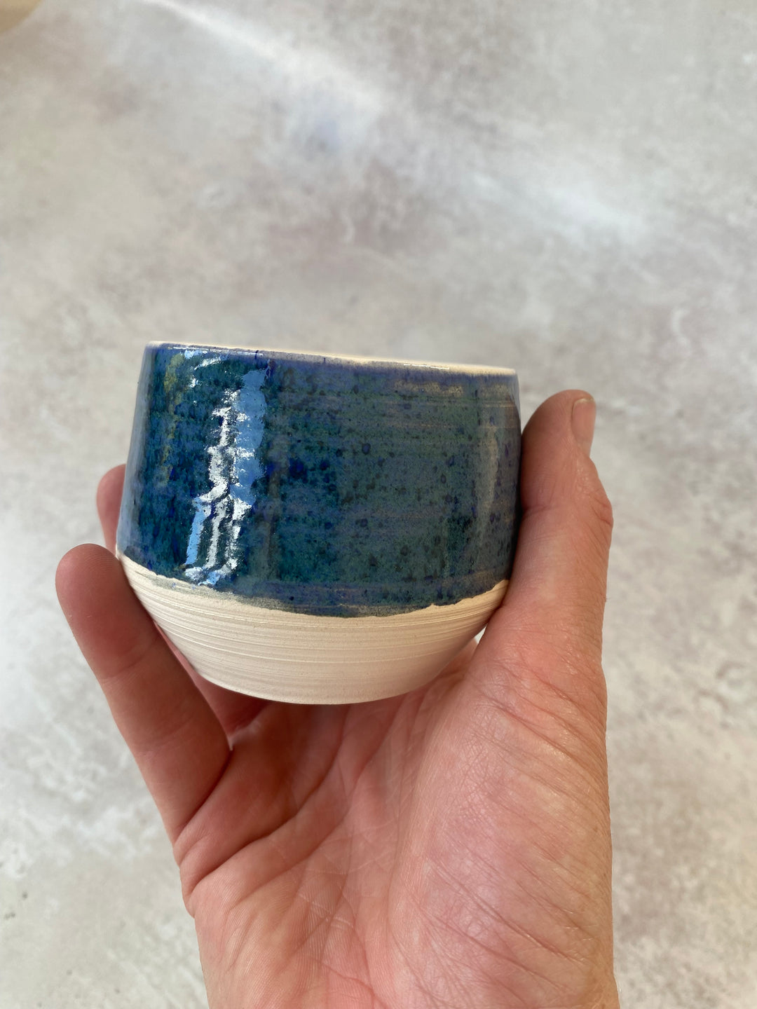 A handmade ceramic cup with a blue glazed top half and a textured unglazed white bottom half, held in a person&