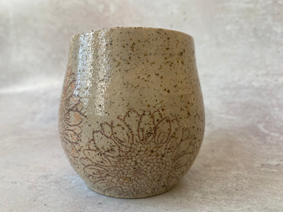 A handmade ceramic cup with a rounded shape, featuring a speckled beige and brown glaze and a brown floral pattern on the lower half, set against a neutral textured background.