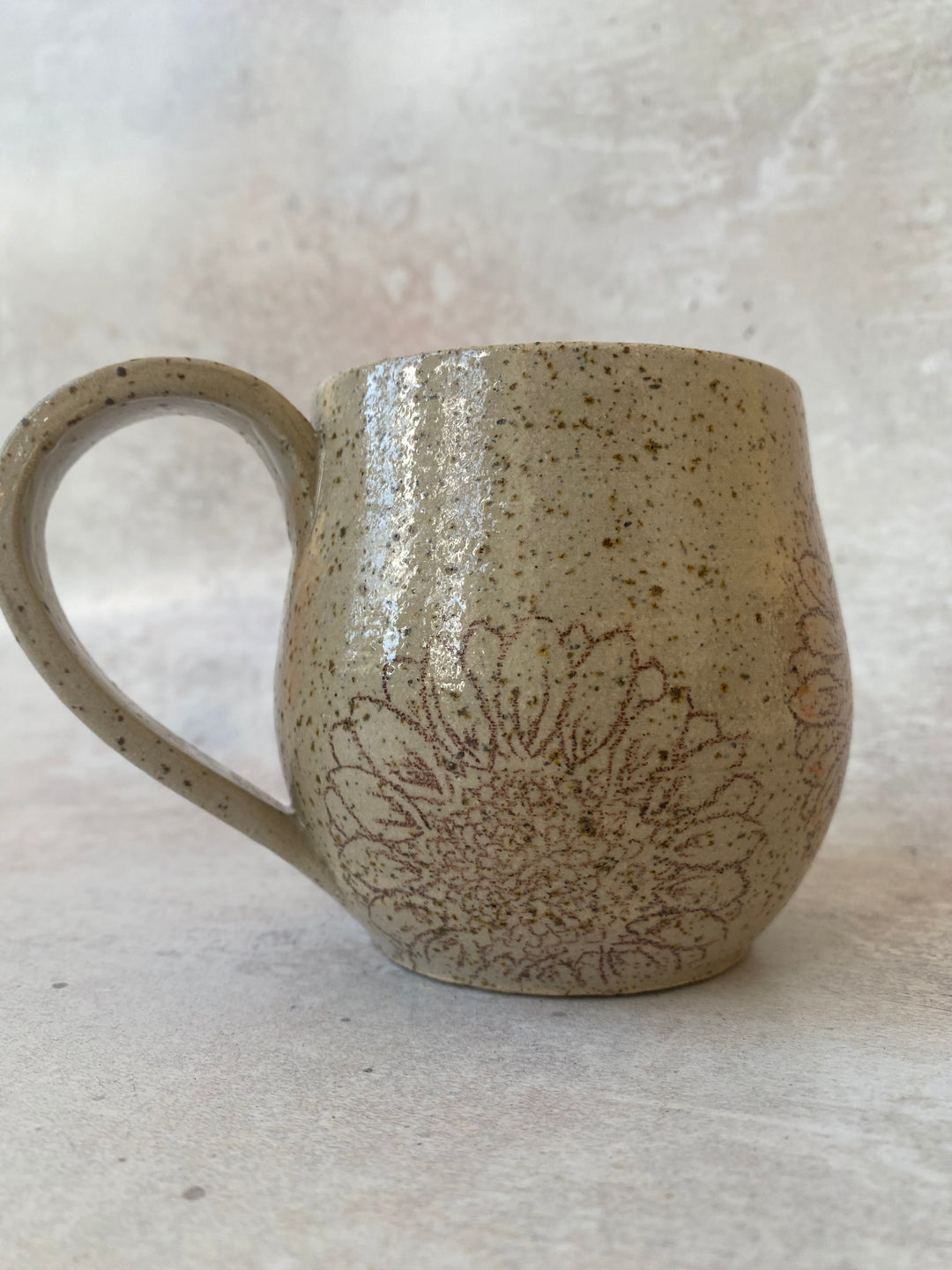 A handmade ceramic mug with a speckled beige and brown glaze, featuring a brown floral line-art pattern on the front, set against a neutral textured background.