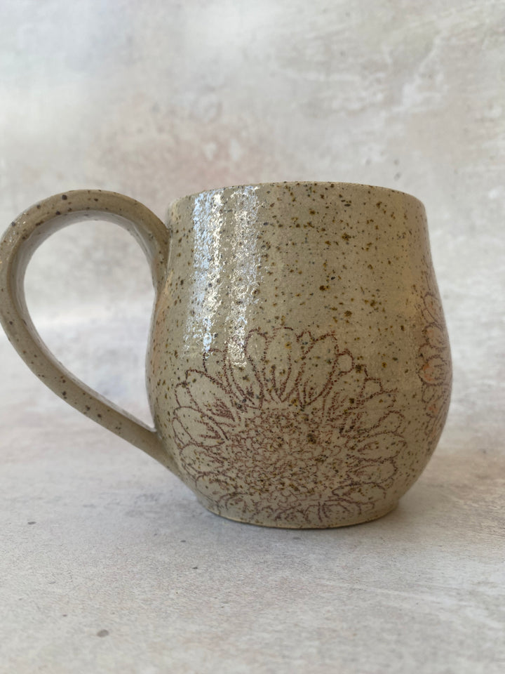 A handmade ceramic mug with a speckled beige and brown glaze, featuring a brown floral line-art pattern on the front, set against a neutral textured background.