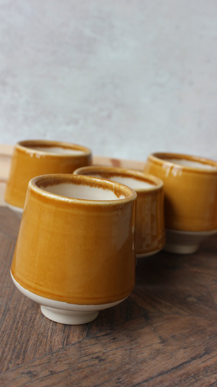 Four handmade ceramic tea cups with a glossy mustard yellow glaze and white unglazed bases, arranged on a dark wooden table against a textured light gray background.