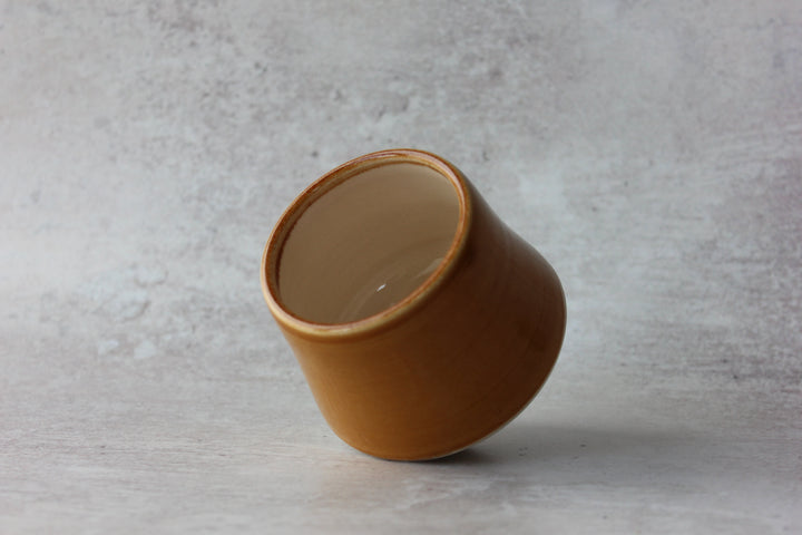 A small tapered ceramic cup with a warm ochre-brown exterior and a lighter beige interior, shown on a textured light gray surface.