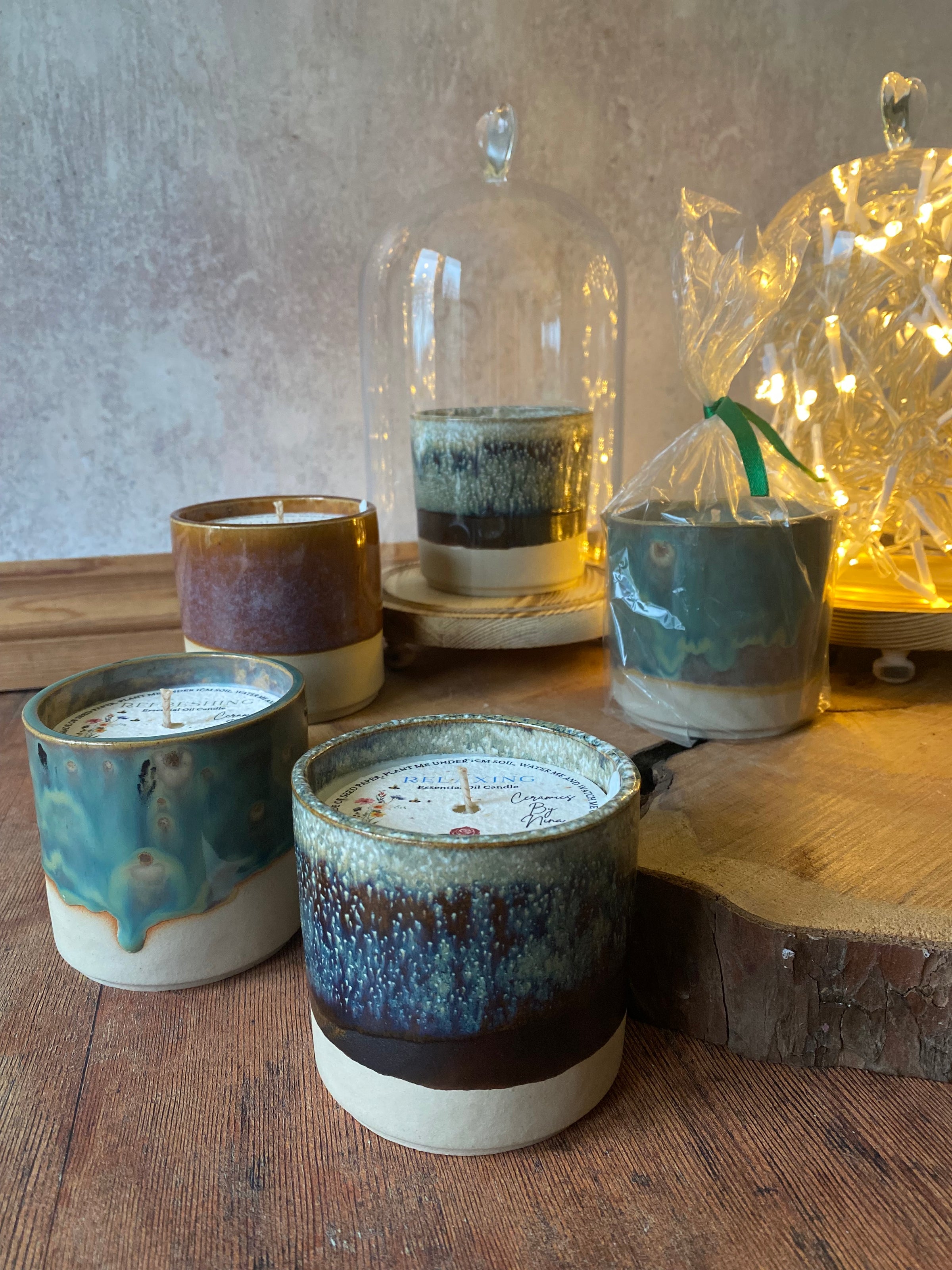 A set of handmade ceramic candles in various glazed finishes, displayed on a rustic wooden table with a glass cloche and warm fairy lights in the background.