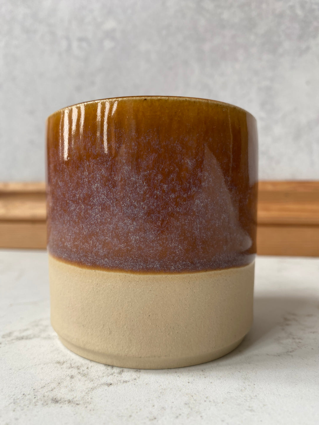 A ceramic wall sconce with a two-tone design, featuring a matte cream-colored base and a glossy amber-brown glazed upper section, mounted on a textured light gray wall.