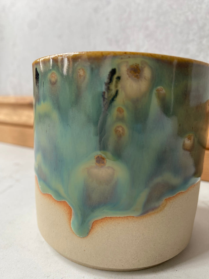 A handmade ceramic cup with a glossy teal and green glaze over a matte beige clay body, featuring a dripping glaze effect and small brown speckles.