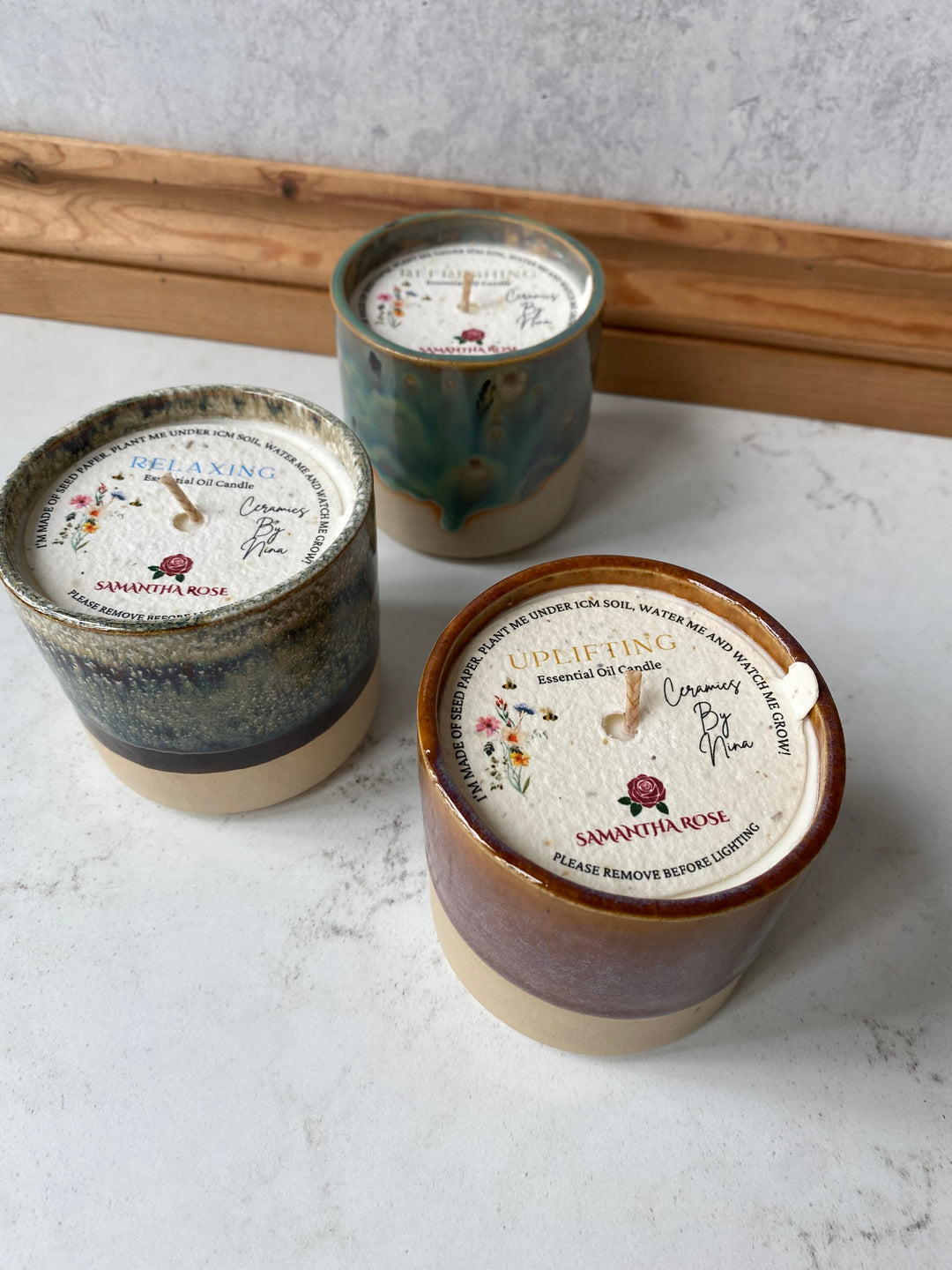 Three ceramic candles with white labels and gold-toned rims, arranged on a light marble surface next to wooden boards. The labels read &