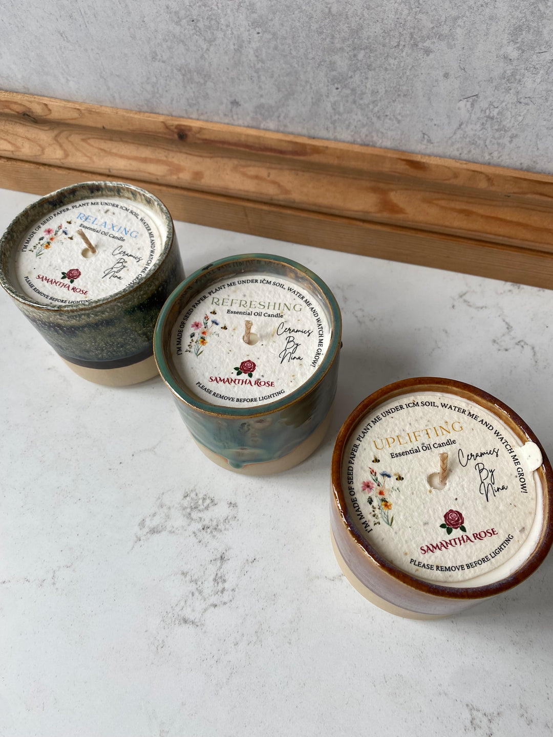 Three small round ceramic jars with decorative lids arranged diagonally on a white marble surface next to a wooden board. The lids feature floral illustrations and text including &