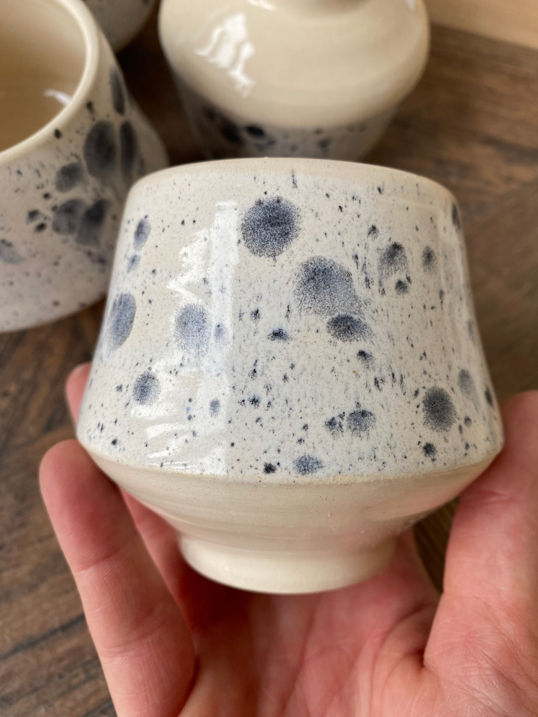 A handmade ceramic cup with a white speckled glaze and dark blue-gray spots, held in a person&