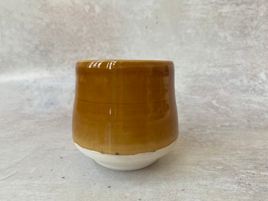 A handmade ceramic cup with a glossy mustard-brown glazed upper half and a matte off-white unglazed base, set against a textured light gray background.