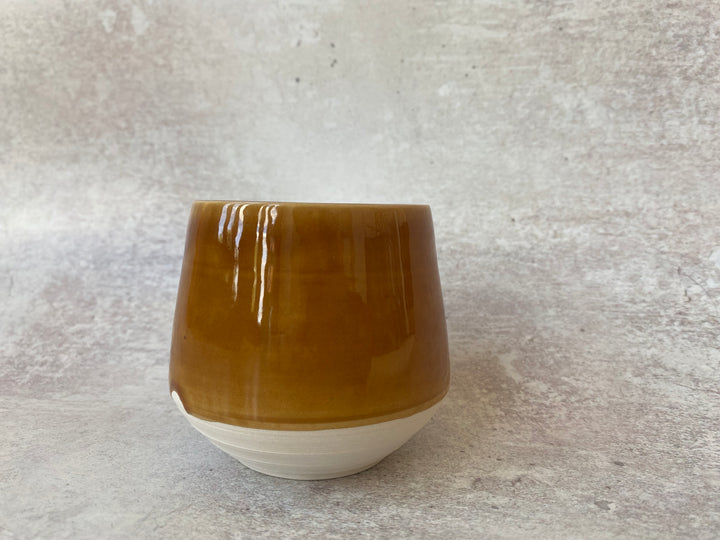 A small ceramic cup with a glossy amber-brown upper body and a matte off-white base, set against a textured light gray background.