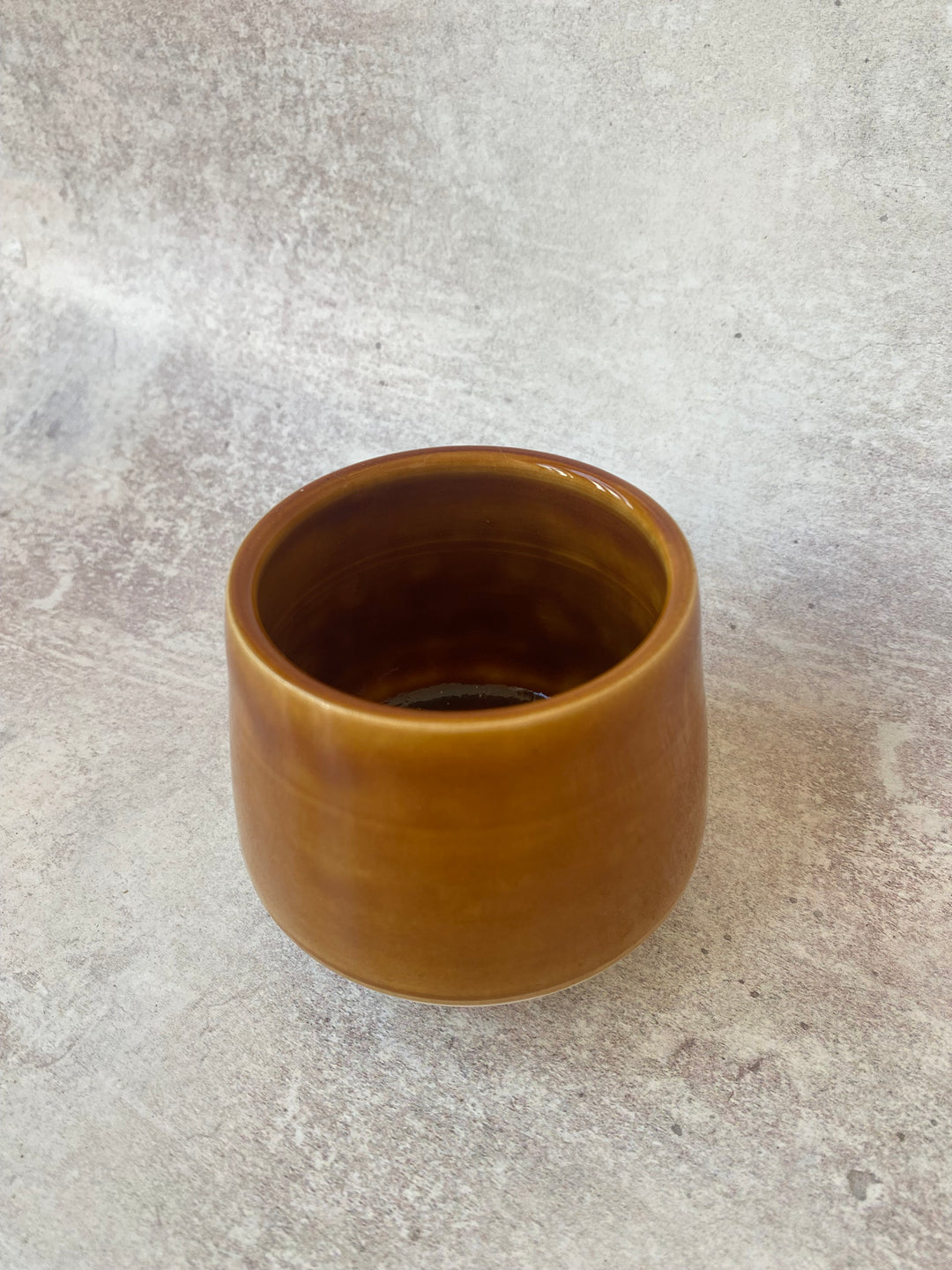 A small, round ceramic cup with a glossy amber-brown finish, set against a textured gray concrete background.