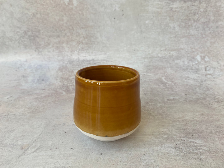A small handmade ceramic cup with a glossy amber-brown glazed finish and a contrasting unglazed white base, set against a textured gray concrete background.