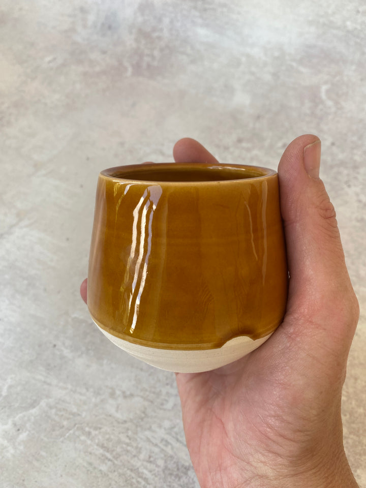 A hand holding a small handmade ceramic cup with a glossy mustard-brown glazed finish and a contrasting unglazed cream-colored base, set against a neutral textured gray background.