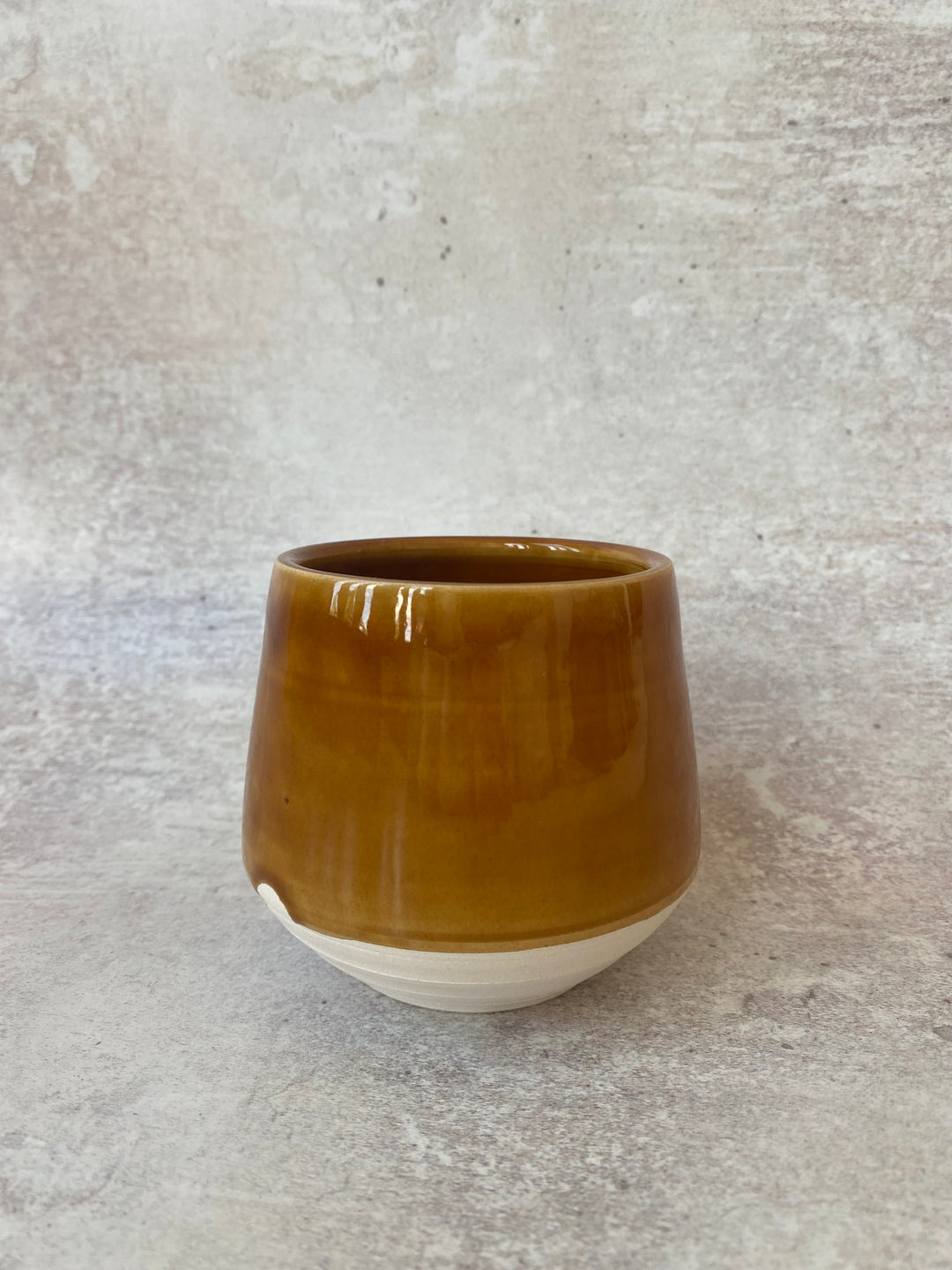 A handmade ceramic cup with a glossy amber-brown glazed upper body and a matte off-white unglazed base, set against a textured gray concrete background.