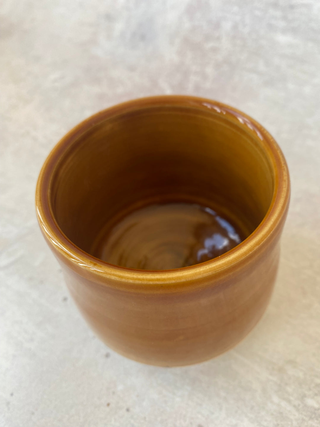 A small handmade ceramic cup with a glossy, warm amber-brown glaze, shown from a top-down perspective on a light textured surface.