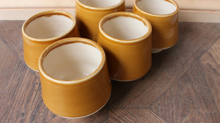 Five small cylindrical ceramic cups with a mustard yellow exterior and cream-colored interiors, arranged on a dark wooden board.