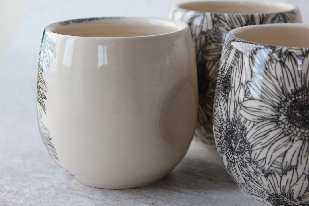 Three ceramic cups with a glossy finish, featuring a black floral sunflower pattern on one cup and a solid cream color on the others, arranged on a light gray surface.