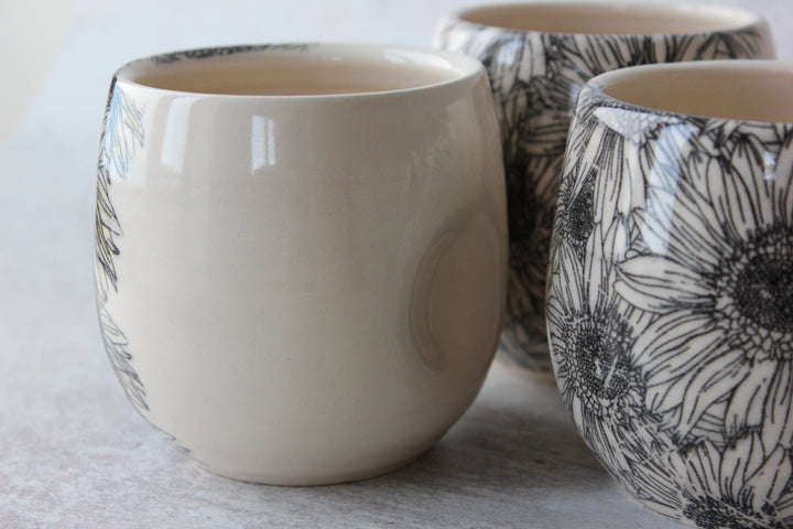Three ceramic cups with a glossy finish, featuring a black floral sunflower pattern on one cup and a solid cream color on the others, arranged on a light gray surface.